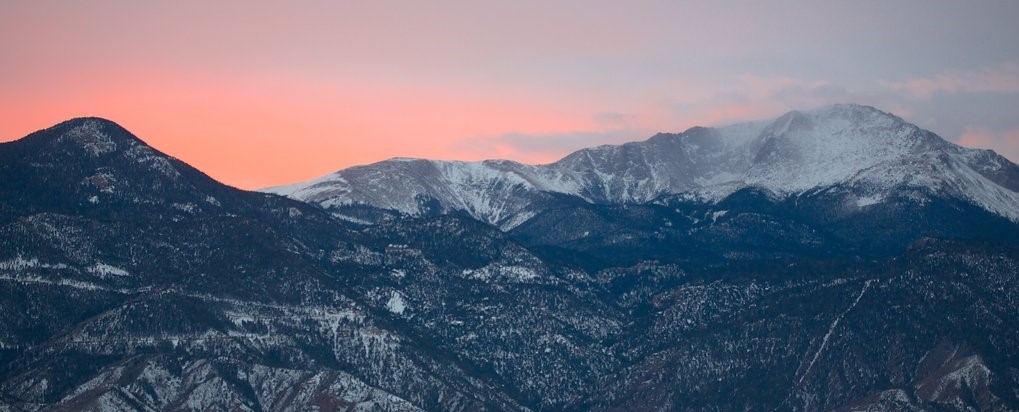 Beautiful picture of a mountain range at sunset. The sky is pink and orange and the mountains are shadowy and have slight snow at the top.
